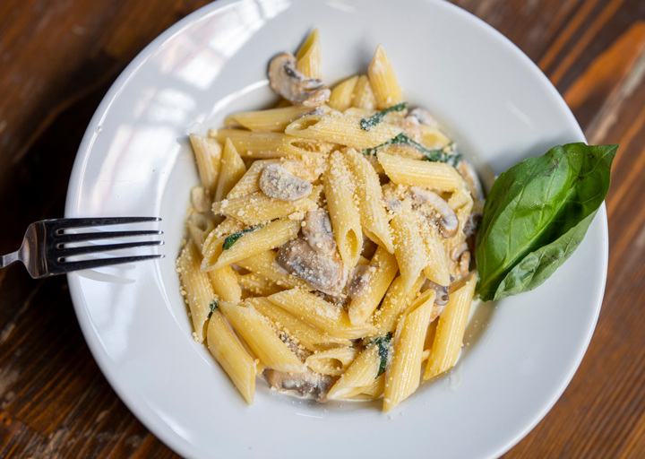 A top-down view of a white plate filled with penne pasta, mushrooms, and green herbs on a dark wooden table, with a fork on the left and a basil leaf on the right.
