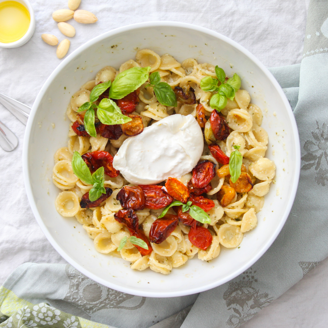 A top-down view of a white bowl filled with orecchiette pasta, topped with a dollop of white sauce, roasted red peppers, and fresh basil leaves, set on a light gray patterned cloth with almonds and a small dish of oil nearby.
