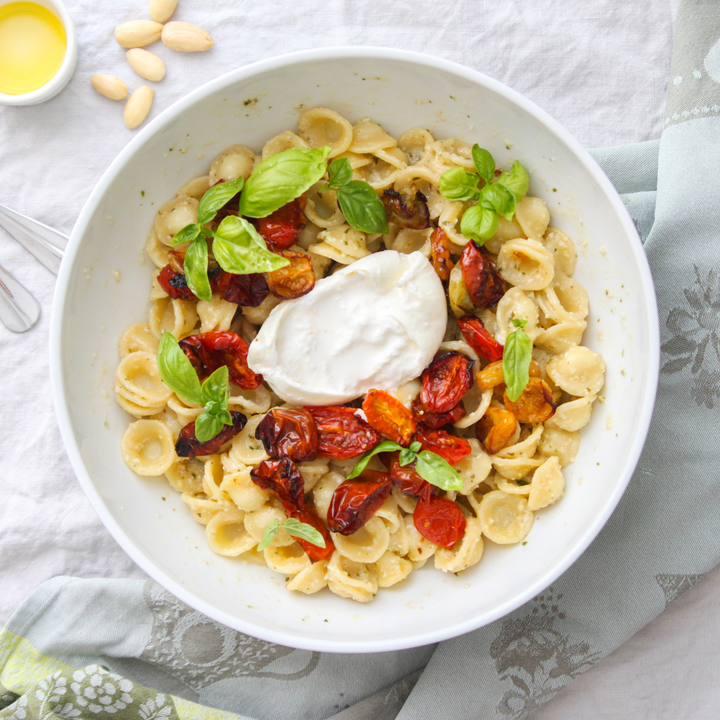 A top-down view of a white bowl filled with orecchiette pasta, topped with a dollop of white sauce, roasted red peppers, and fresh basil leaves, set on a light gray patterned cloth with almonds and a small dish of oil nearby.