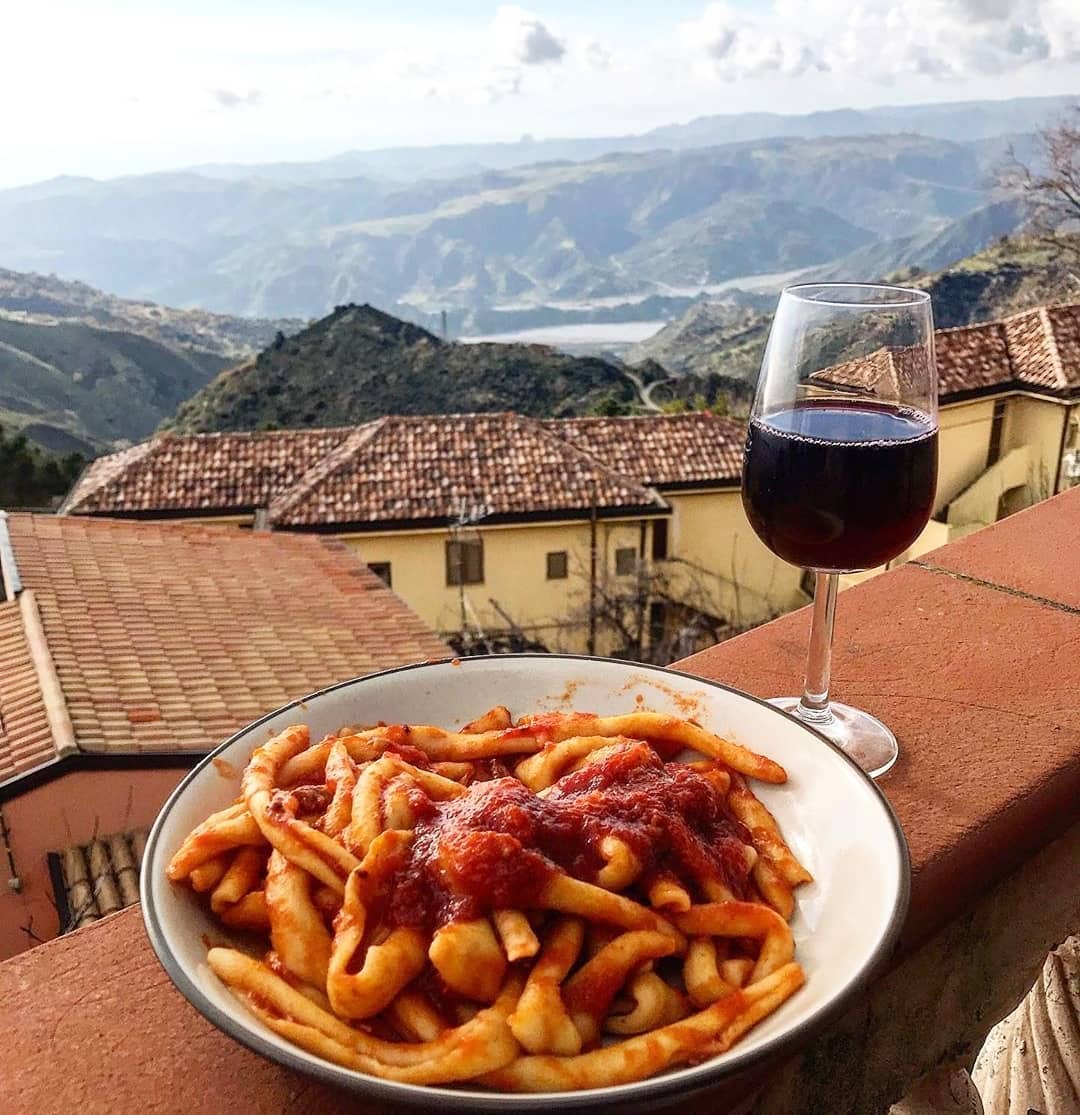 A bowl of pasta with red sauce sits on a balcony next to a glass of red wine, with a scenic mountain and village view in the background.