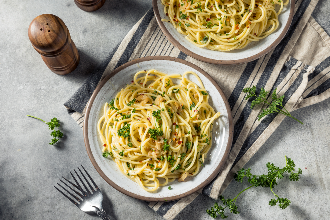 Two plates of creamy pasta with herbs and small orange garnish, arranged on a gray stone surface with a striped napkin, fork, pepper mill, and parsley sprigs.