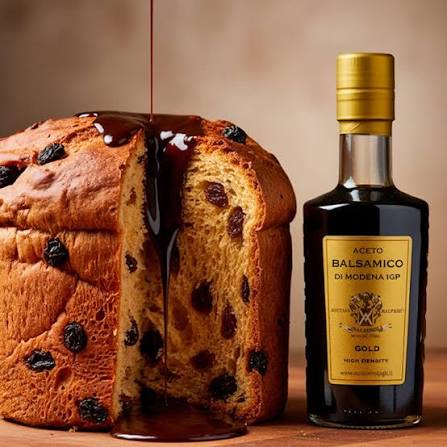 A sliced panettone cake  with dark dried fruit pieces and a glossy drizzle of dark syrup, shown next to a bottle of Modena IGP Gold Balsamic Vinegar on a wooden surface against a warm beige background.