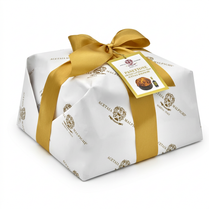 A gift-wrapped panettone from Acetaia Malpighi, wrapped in white paper with gold foil patterns and tied with a large gold ribbon bow, on a white background.
