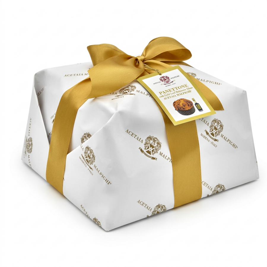 A gift-wrapped panettone from Acetaia Malpighi, wrapped in white paper with gold foil patterns and tied with a large gold ribbon bow, on a white background.