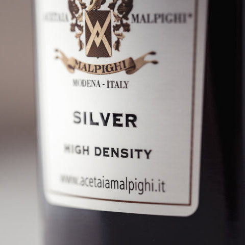 Close-up of a bottle label for Acetaia Malpighi Silver High Density, showing the brand crest and text on a dark bottle against a blurred background.