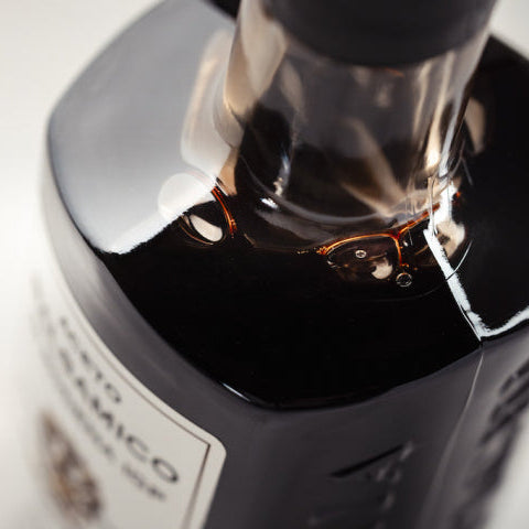 Close-up of a dark glass bottle with a white label and a black cap, photographed on a light neutral background with shallow depth of field.