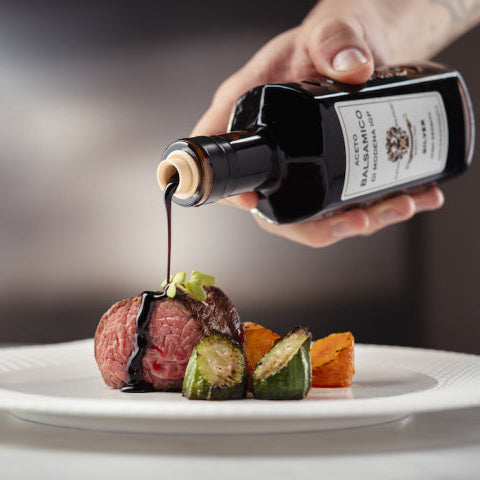 Person pouring dark sauce from a bottle onto a dish of food on a white plate.