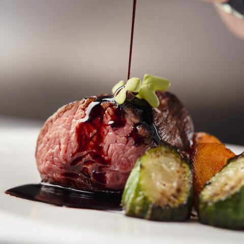 A plated gourmet dish featuring a medium-rare beef steak topped with a dark sauce, garnished with a small green herb sprig, served with zucchini and roasted sweet potato wedges on a white plate.