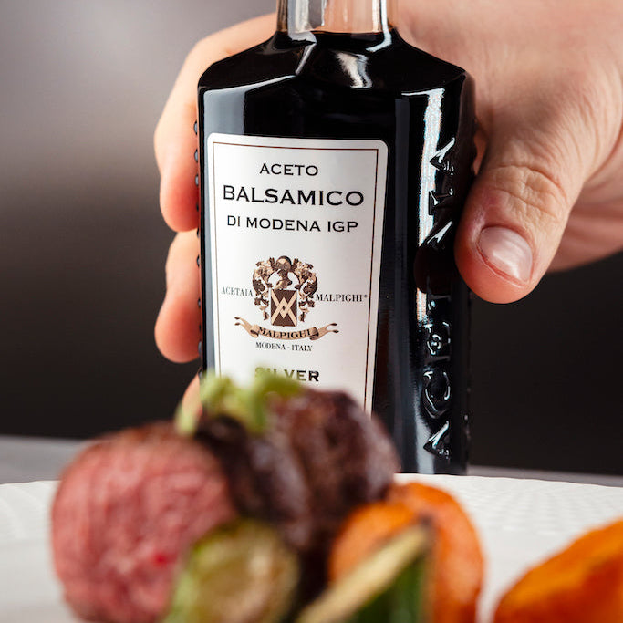 A hand holding a bottle of Modena IGP balsamic vinegar, with blurred food items on a plate in the foreground against a dark gray background.