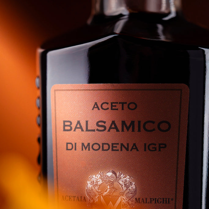 A close-up of a dark glass bottle labeled 'ACETO BALSAMICO DI MODENA IGP' with a warm copper-brown label, photographed against a blurred orange background.