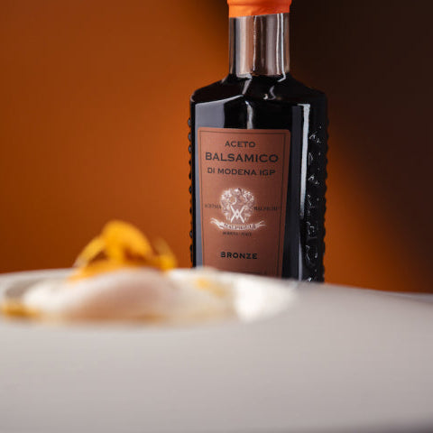 A bottle of Modena balsamic vinegar labeled 'Aceto Balsamico di Modena' stands on a white plate with a small orange garnish against a warm brown background.