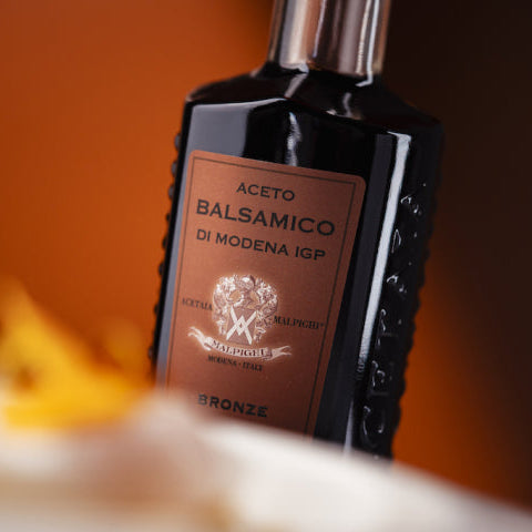 A dark bottle of Modena IGP balsamic vinegar with a bronze label, shown in a close-up shot against a warm orange-brown background with a blurred foreground.