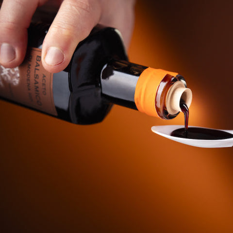 A close-up of a hand holding a dark bottle and pouring a dark liquid, likely balsamic vinegar, into a white spoon held by another hand, against a warm brown background.