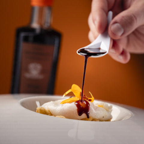 A close-up of a hand holding a spoon and pouring a dark syrup or sauce over a plated dessert, with a blurred bottle in the background against a warm orange backdrop.