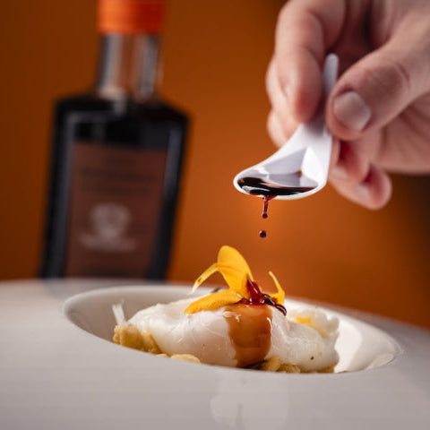 A close-up of a hand holding a small white spoon and drizzling a dark liquid over a plated food dish, with a blurred bottle in the background against a warm orange backdrop.
