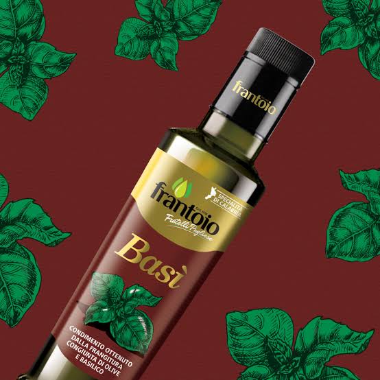 A bottle of Frantoio Basí olive oil, shown diagonally against a dark red background decorated with green basil leaf illustrations. The bottle features a black cap and a label with gold and brown sections.