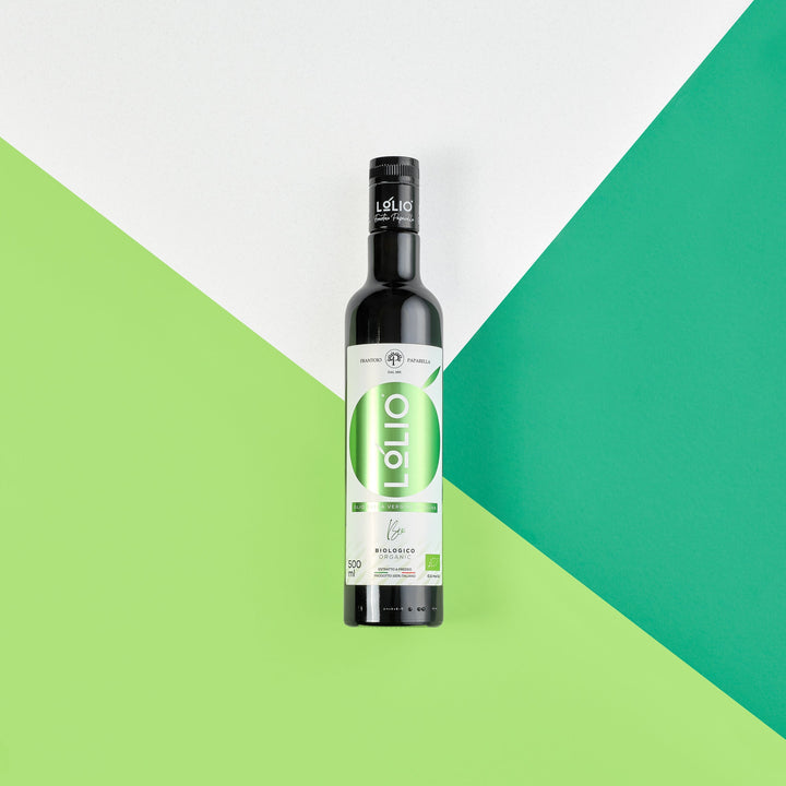 A bottle of olive oil stands upright on a geometric background split diagonally into white, green, and teal sections. The bottle is dark glass with a black cap and a white label featuring a large green oval and the word 'olio' in white.