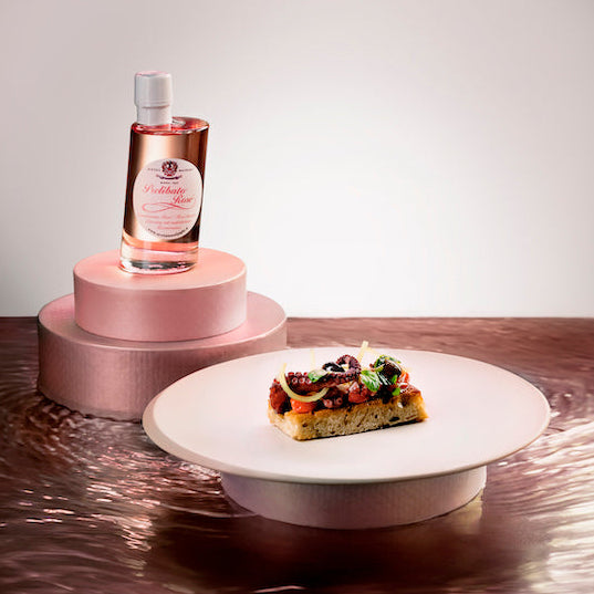A bottle of Prelibato Rose Dressing by Acetaia Malpighi sits on a pink tiered pedestal, while a small slice of gourmet pizza with herbs rests on a white plate in the foreground, all set on a reflective metallic surface against a neutral studio background.
