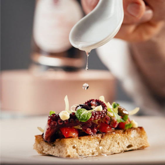 A close-up of a hand holding a white ceramic spoon and drizzling oil over a slice of toasted bread topped with red fruit, green herbs, and white cheese shavings, with a blurred bottle in the background.