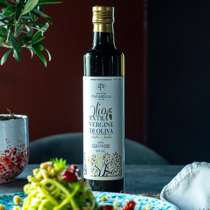 A bottle of Paparella d'Olio extra virgin olive oil stands on a table in a styled kitchen or dining setting, with a plate of pasta in the foreground and greenery in the background.