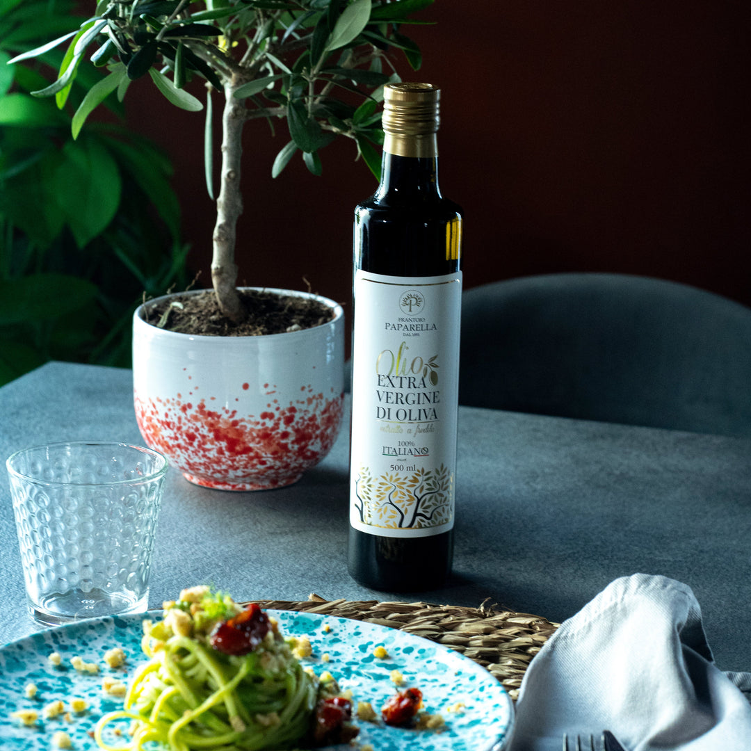 A bottle of olive oil stands on a dining table beside a plate of pasta, a glass, cutlery, and a potted olive tree against a dark background.