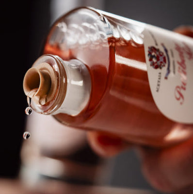 A close-up shot of a hand holding a clear glass bottle filled with amber liquid, with a single drop of liquid dripping from the open bottle against a dark background.