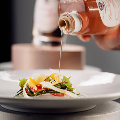 A close-up of a hand pouring a clear liquid from a glass bottle onto a plated salad, with a dark background and a blurred bottle behind the plate.