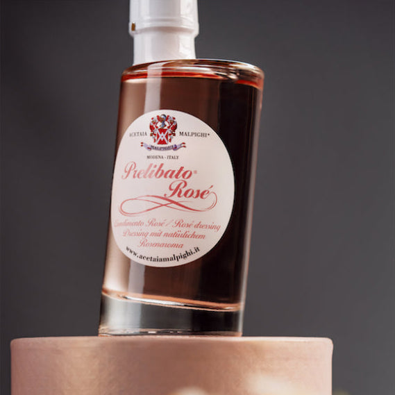 A bottle of Prelibato Rose Dressing Acetaia Malpighi standing upright on a round pedestal, with a blurred foreground and dark gray background.