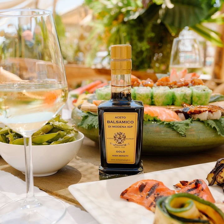 A bottle of Aceto Balsamico di Modena Gold balsamic vinegar stands on a table surrounded by plated food, including sushi, grilled salmon, green beans, and a glass of white wine, with a blurred outdoor dining background.