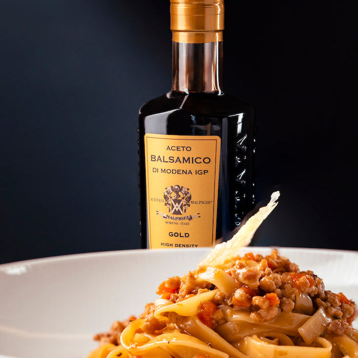 A bottle of Modena ICP Balsamic Vinegar with a gold cap and label stands behind a white bowl of pasta with meat sauce, with a toothpick holding a slice of bread.