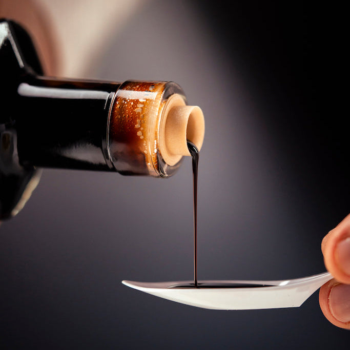 A close-up of a dark bottle pouring a thick, dark brown liquid, likely balsamic vinegar, in a thin stream onto a white spoon held by a hand against a dark background.