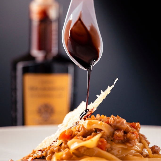 A close-up of a plated pasta dish with meat sauce, parmesan cheese, and a hand drizzling dark balsamic syrup from a white spoon, with a bottle in the blurred background.