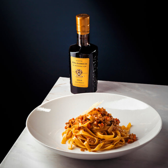 A white plate of pasta with a meat sauce sits on a light marble surface, with a dark glass bottle featuring a gold cap and yellow label standing behind it against a dark background.