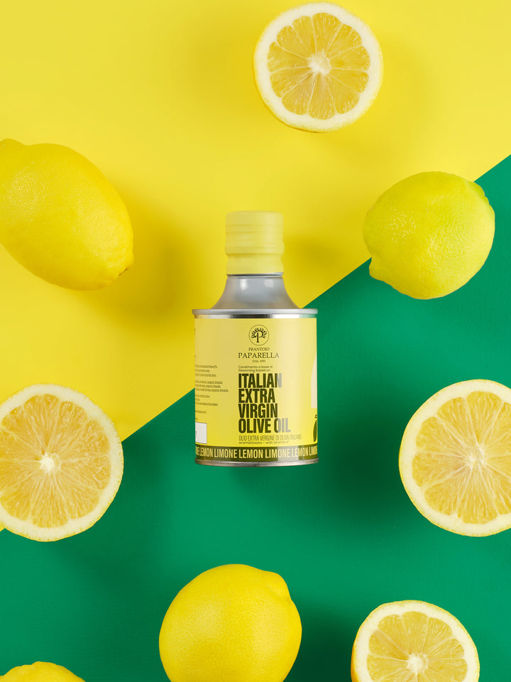 A bottle of Papiarella Italian Extra Virgin Olive Oil centered on a bright yellow and green background, surrounded by whole and sliced lemons.