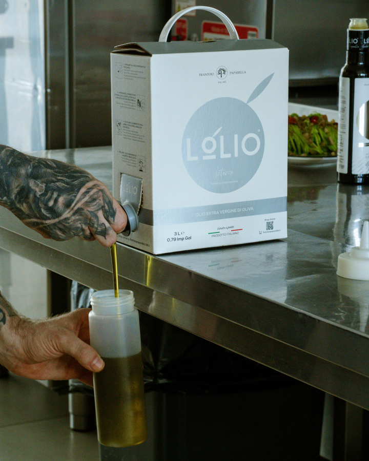 A tattooed hand pours olive oil from a Lólio branded cardboard dispenser box into a clear plastic bottle on a stainless steel kitchen counter.