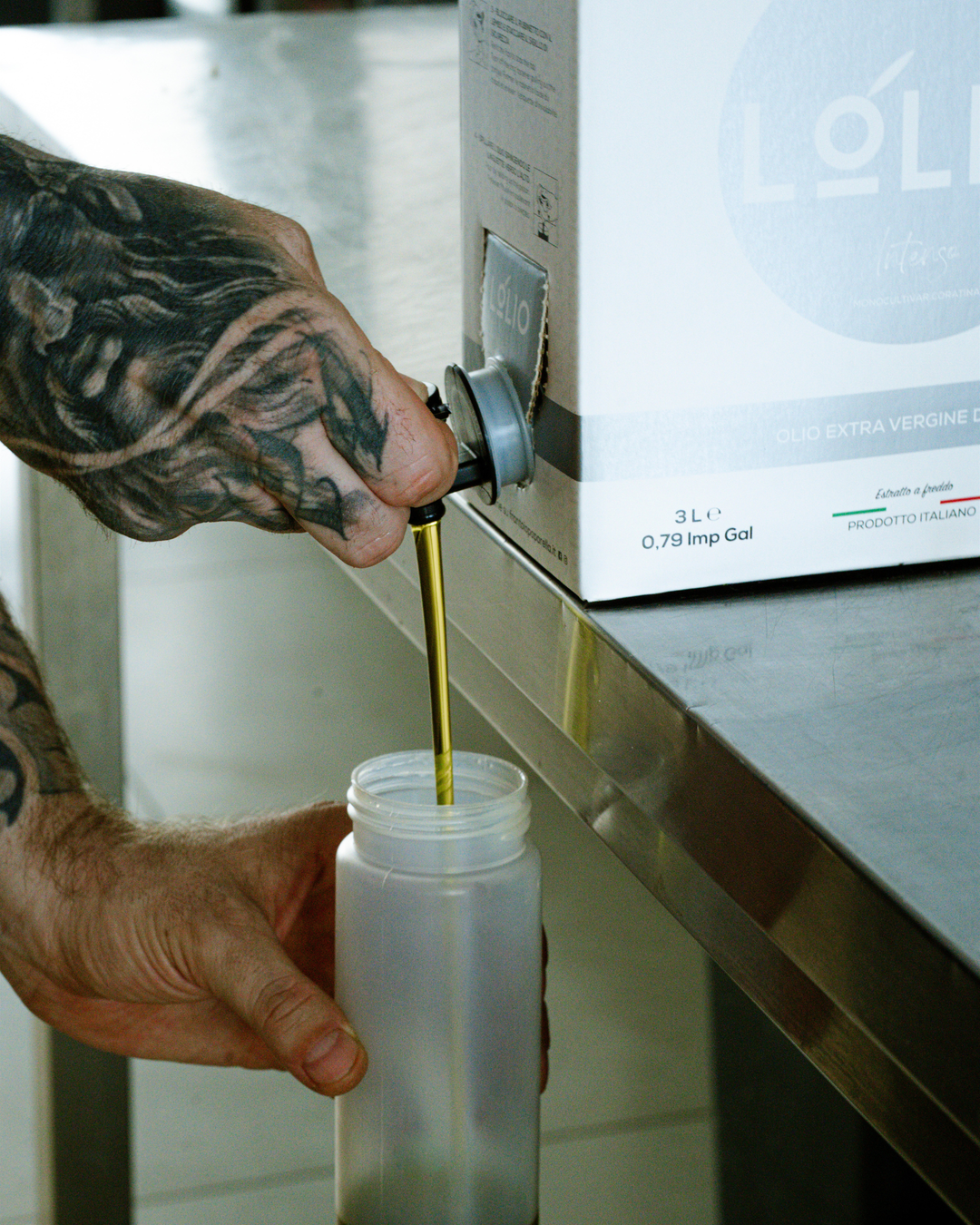 A tattooed hand holds a metal spout over a clear plastic bottle, pouring a golden extra virgin olive oil. A white box labeled LOLIO is visible on a stainless steel counter in the background.