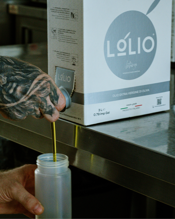A tattooed hand dispenses olive oil from a Lólio branded cardboard dispenser into a translucent plastic bottle on a stainless steel counter.