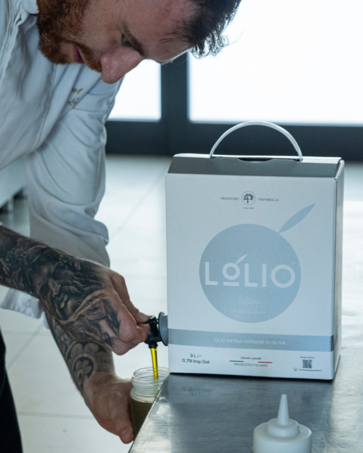 A man with a tattooed arm pours olive oil from a large white Lolio box into a small bottle on a stainless steel counter.