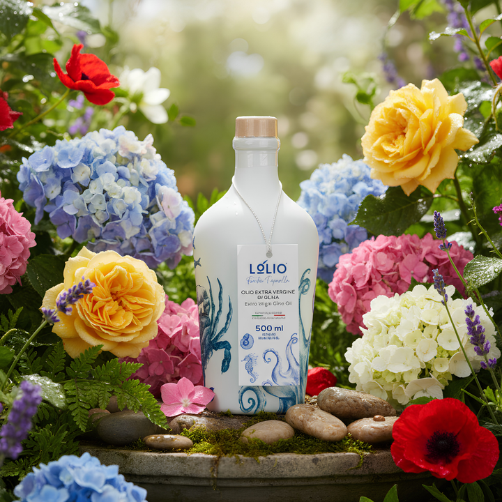 A white glass bottle of Lólio extra virgin olive oil with blue wave illustrations and a gold cap, surrounded by a lush garden arrangement of blue, pink, yellow, and red flowers, green leaves, and small stones.