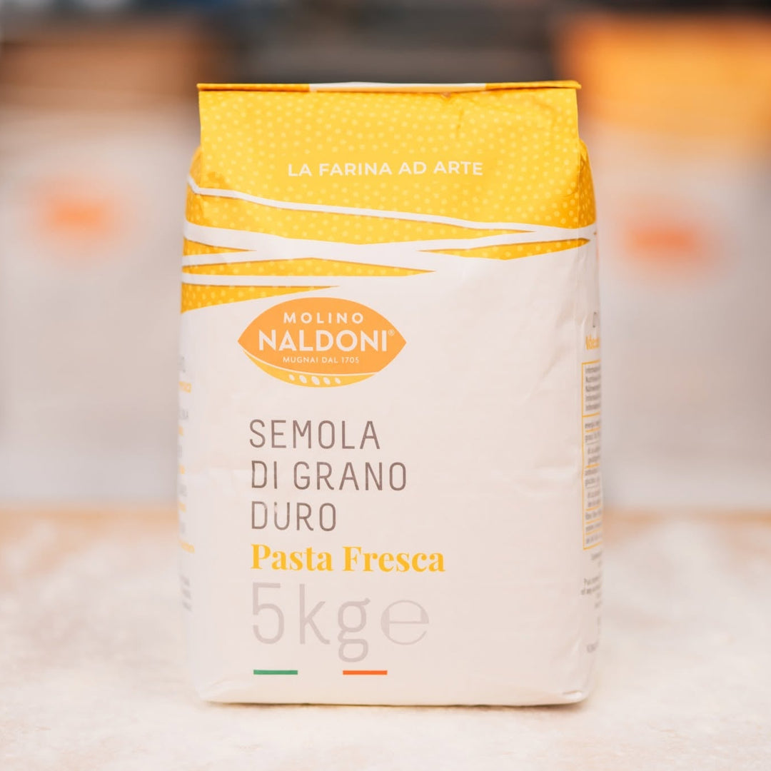 A 5kg bag of Molino Naldoni Semola di Grano Duro Pasta Fresca durum wheat flour, standing on a floured surface with a blurred bakery background.