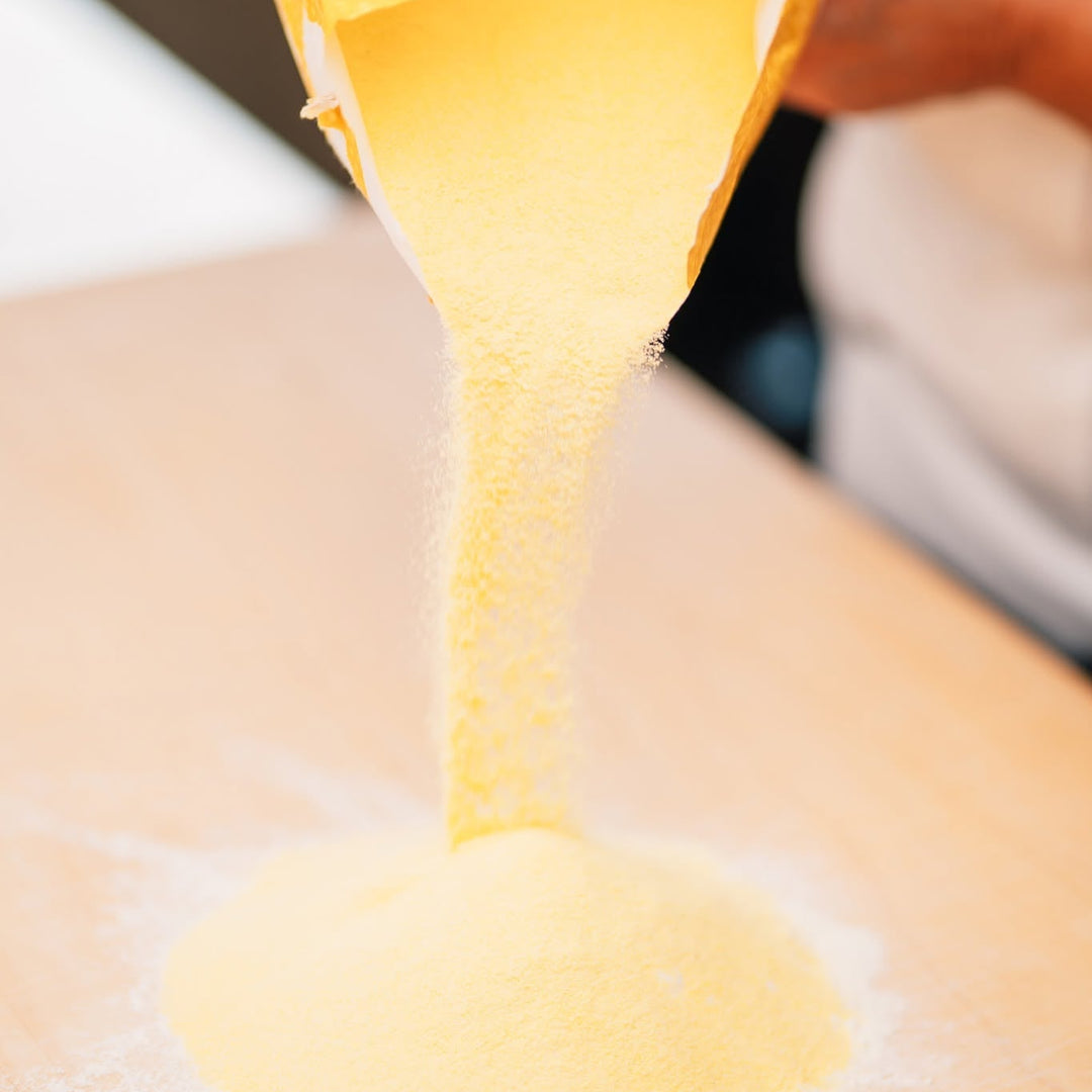 Yellow flour, being poured from a yellow paper bag onto a light wooden surface.