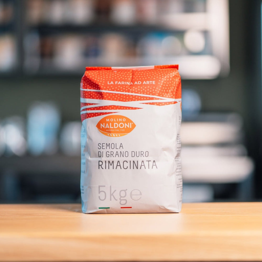 A 5kg bag of Molino Naldoni Semola di Grano Duro Rimacinata durum wheat flour, standing on a wooden table with a blurred kitchen background.