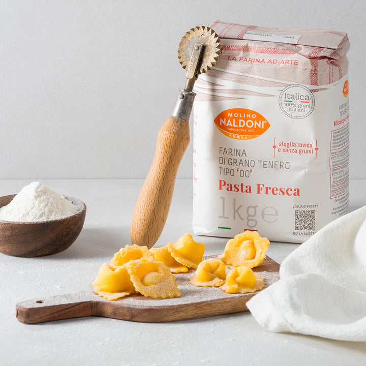 A bag of Molino Naldoni Pasta Fresca durum wheat flour stands on a light surface next to a wooden rolling pin, cut yellow pasta on a wooden board, a small bowl of white flour, and a white cloth.