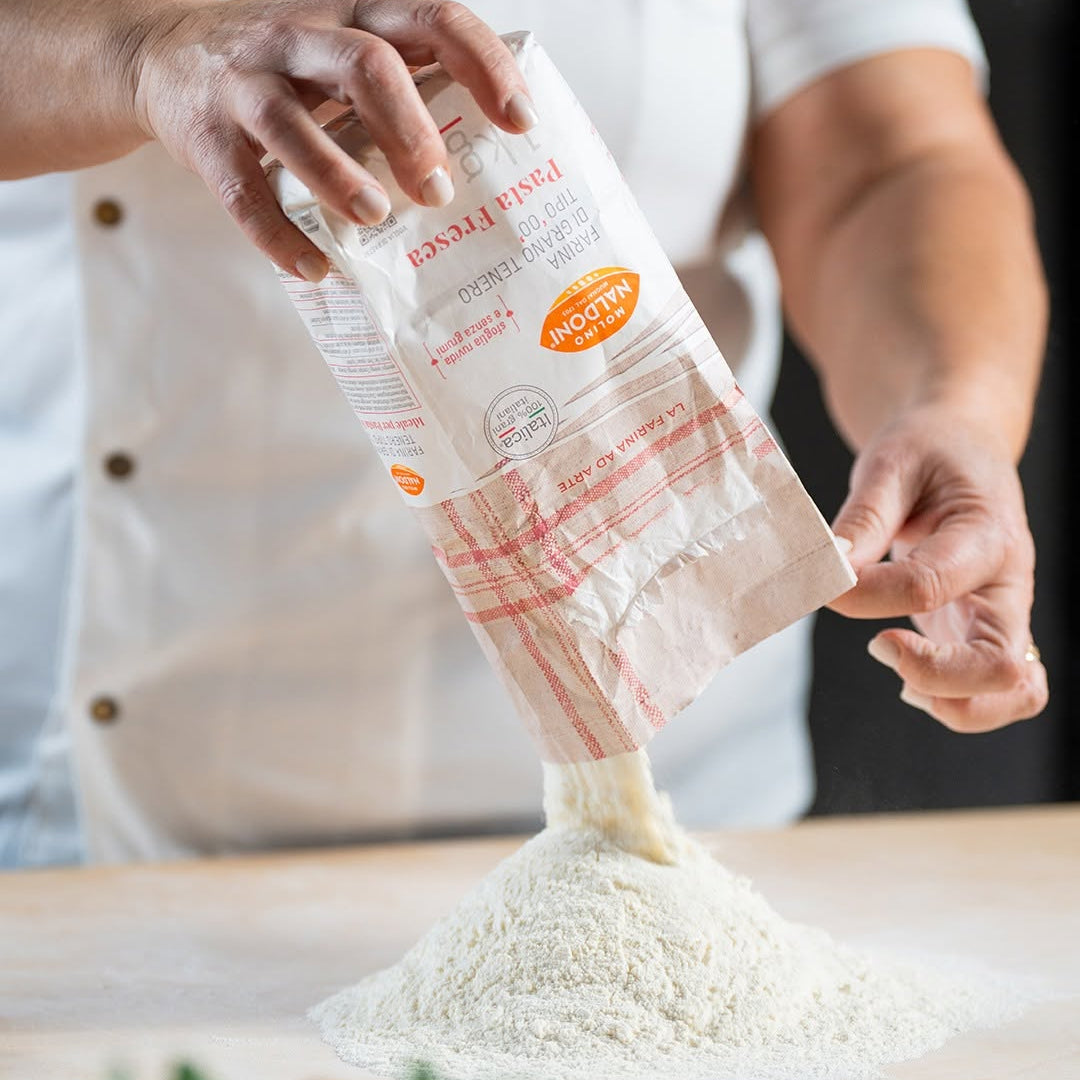 A person in a white chef's coat pours flour from a paper bag onto a wooden surface. The bag has red stripes and an orange circular label that reads 'MEDIUM'.