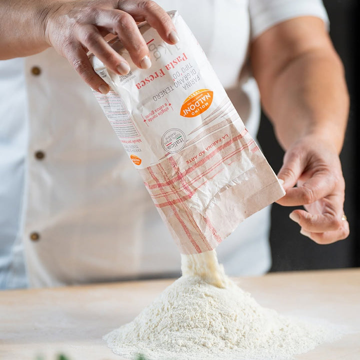 A person in a white chef's coat pours flour from a paper bag onto a wooden surface. The bag has red stripes and an orange circular label that reads 'MEDIUM'.
