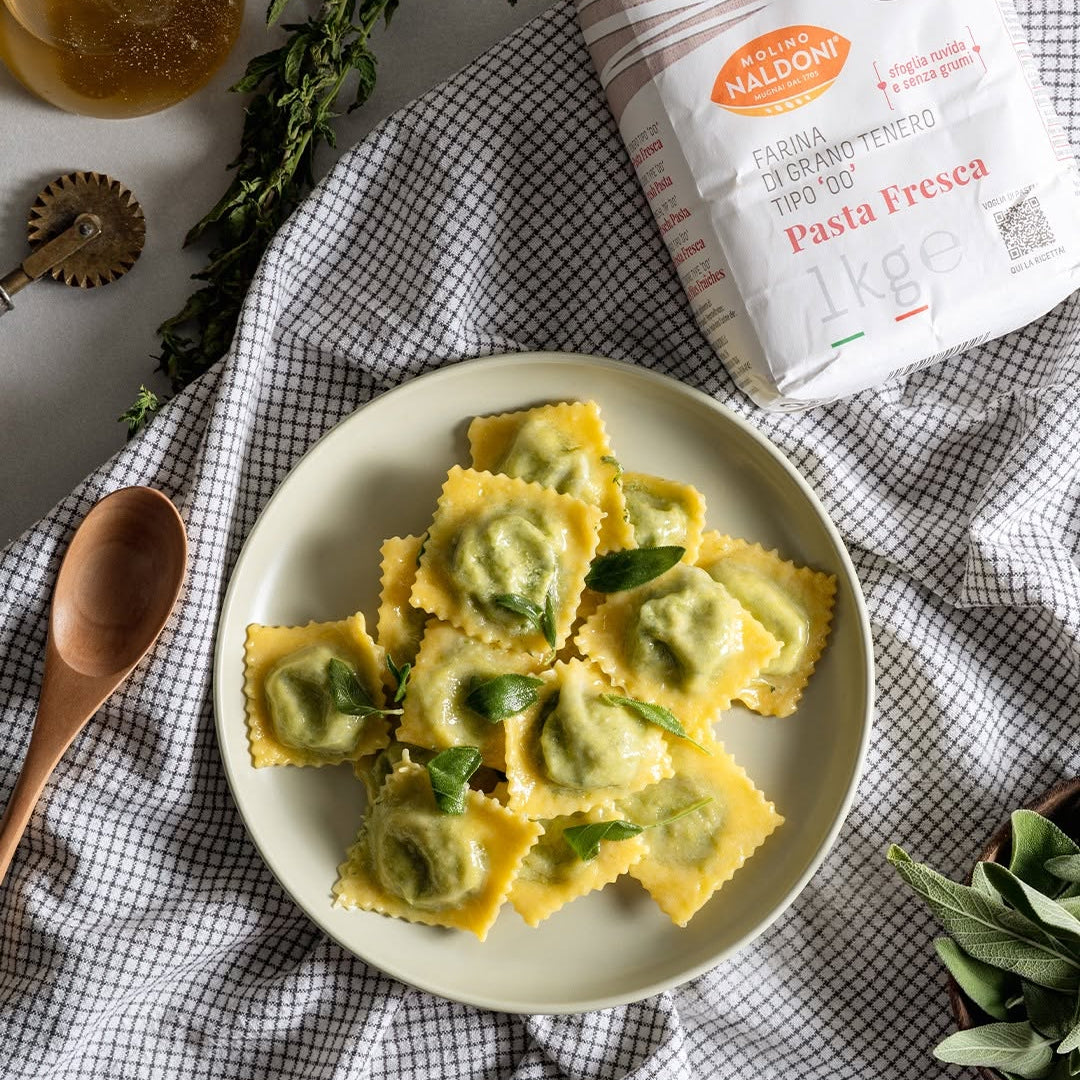A plate of fresh handmade pasta with green sauce, styled with a bag of Naldoni flour, a wooden spoon, and herbs on a checkered cloth.