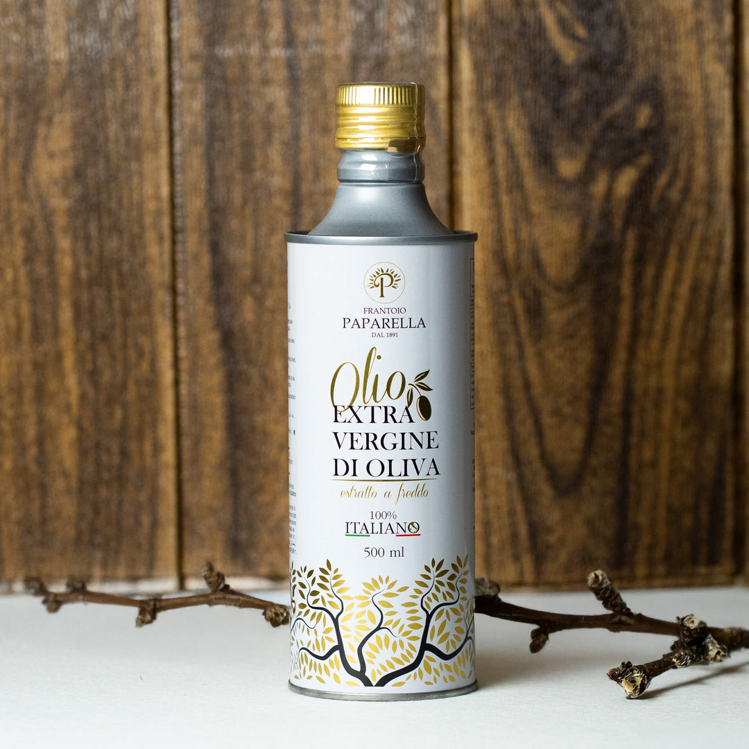 A 500ml bottle of Paparella Extra Vergine di Oliva oil with a gold cap and a white label decorated with gold olive tree illustrations, set on a white surface with rustic wooden branches and green olives against a wooden background.
