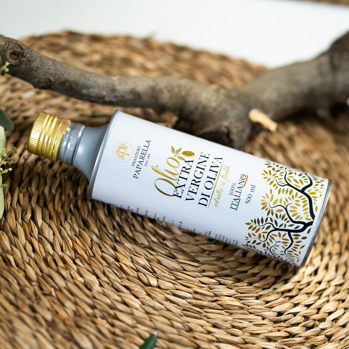 A 500 ml bottle of Paparella Extra Vergine d'Olio olive oil, featuring a white label with gold and black text and graphics, resting on a woven straw mat with a rustic branch in the background.
