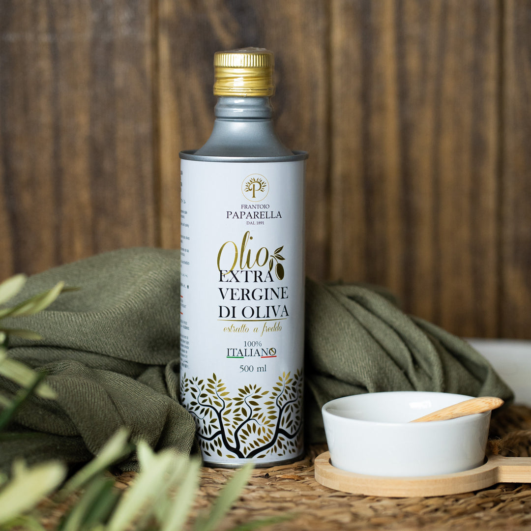 A bottle of Paparella Extra Vergine d'Olio olive oil stands on a woven tray beside a small white bowl with a wooden spoon, with olive branches and green olives in the foreground against a rustic wooden background.