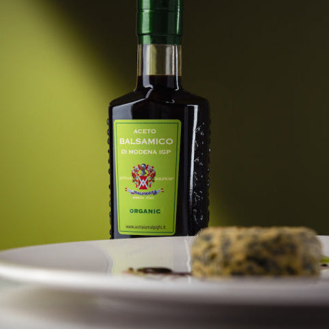 A bottle of Modena IGP organic balsamic vinegar stands on a white plate with a piece of bread in front, set against a dark olive-green background.